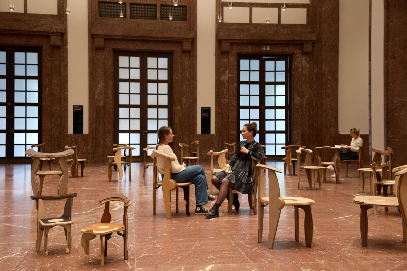 Viele hölzerne Stühle stehen im Raum verteilt. Zwei Frauen sitzen sich gegenüber, eine andere sitzt im Hintergrund.