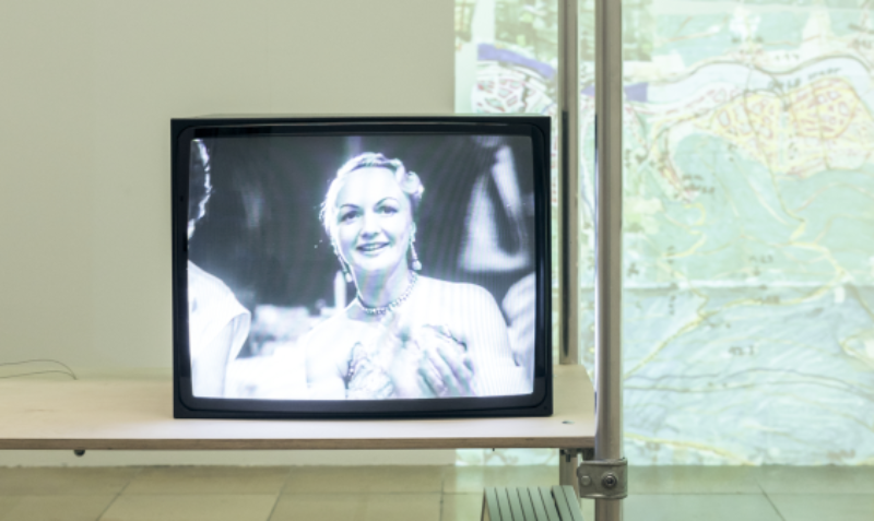 Ein schwarz-weiß Film läuft auf dem Fernseher, eine Frau ist zu sehen. Im Hintergrund eine Projektion einer Landkarte.
