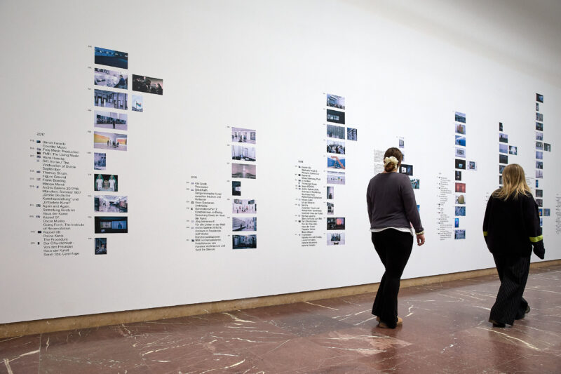 Zwei Personen laufen an einer Wand vorbei an der mehrere kleine Bilder angebracht sind.