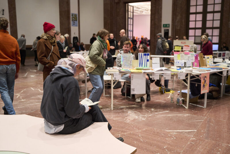 Ein belebter Raum mit Büchertischen und Besucher*innen.