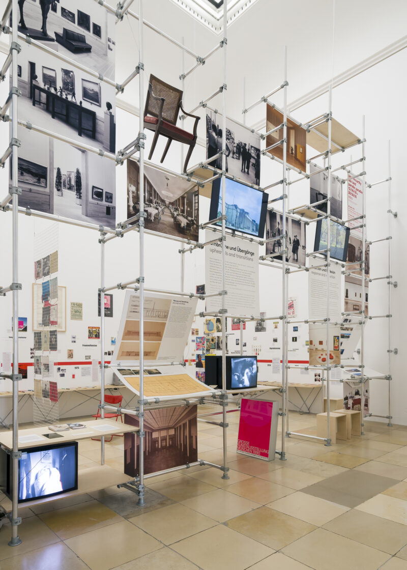 Im Ausstellungsraum ist ein großes Gerüst, an dem Fotos und Gegenstände hängen und Bildschirme angebracht sind, die Videos zeigen.