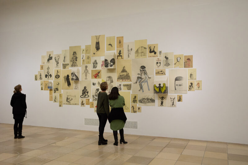 Drei Personen schauen sich an einer Wand kleine Bilder an.