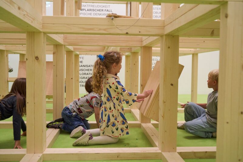 Kinder spielen in einem hölzernen Gerüst auf grünem Boden.