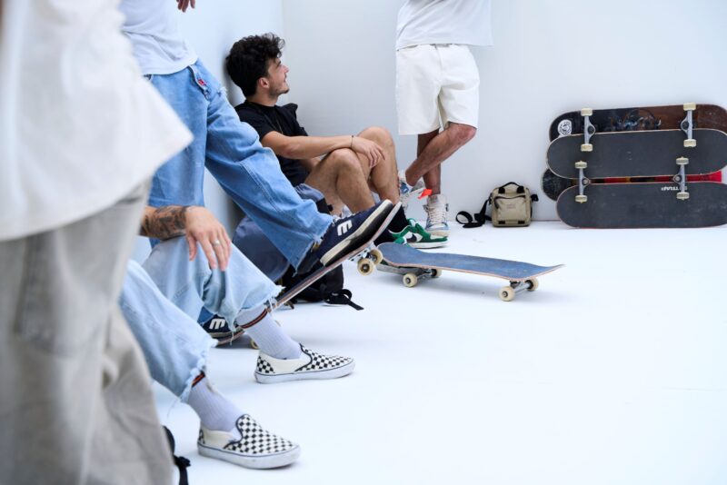 Auf weißem Grund sitzen Personen von denen nur die Füße zu sehen sind. Im Hintergrund befinden sich mehrere Skatboards.