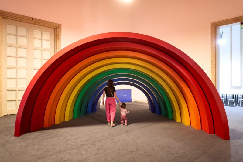 Eine Person geht mit einem Kind unter einem großen bunten Regenbogen durch.