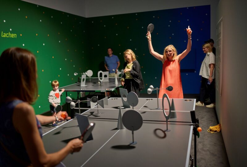 Menschen spielen Tischtennis. Auf der Tischtennisplatte befinden sich abstrakte Skulpturen.