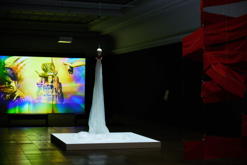 Im dunklen Ausstellungsraum wird im Hintergrund ein farbenfroher Film gezeigt, im Vordergrund eine Skulptur aus rotem Stoff. In der Mitte des Raums schwebt eine Skulptur, ein Kopf mit einem Körper aus langem weißen Stoff.