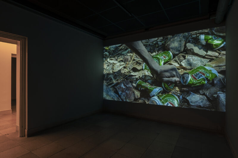 Auf dem Filmstill greift eine Hand auf dem Waldboden nach zerdrückten Getränkedosen.