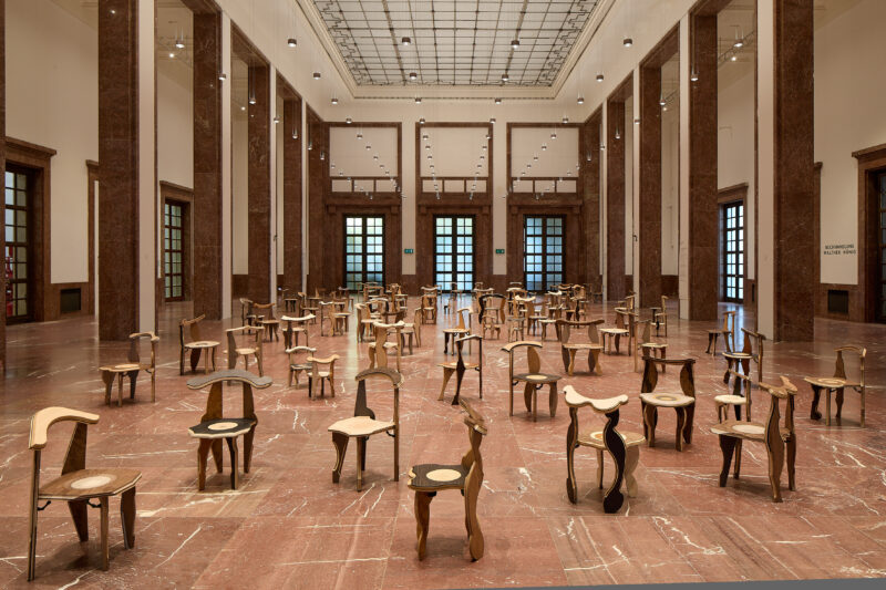 In einer großen Halle stehen zahlreiche individuelle Stühle aus Holz verteilt auf dem rot-braunen Marmorboden.