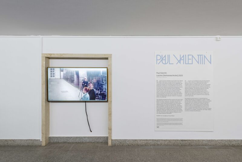 An der weißen Wand ein Bildschirm, auf dem man den Flur mit Personen in Liveaufnahme sieht. Daneben ein Infotext zu der Installation.