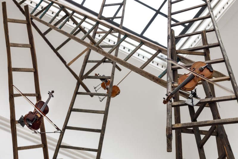 The artwork consists of many assembled wooden ladders to which violins are attached.