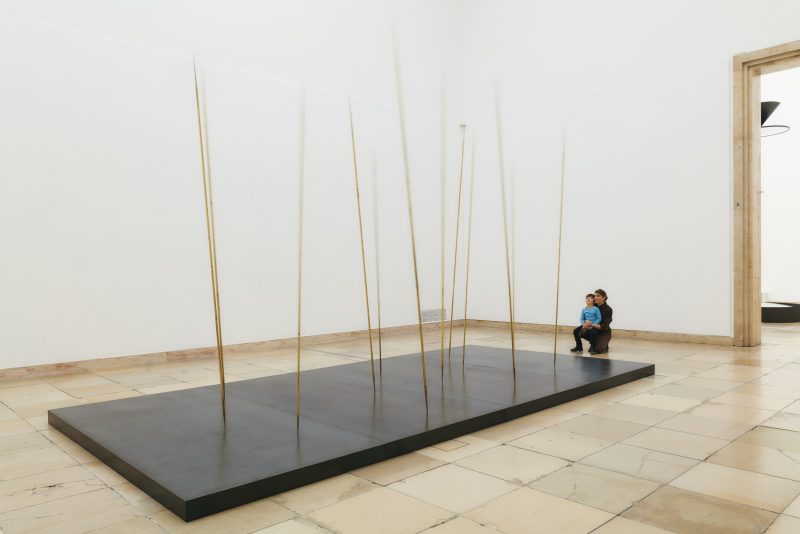 A dark panel on which long brass rods stand at an angle. A mother and child looking at the work.