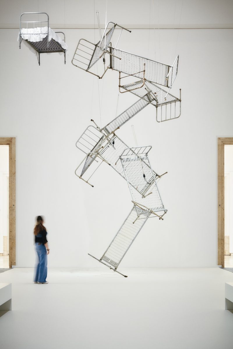 Bed frames are assembled in a spiral and float in the high exhibition space. In front of them is a person looking at the work.