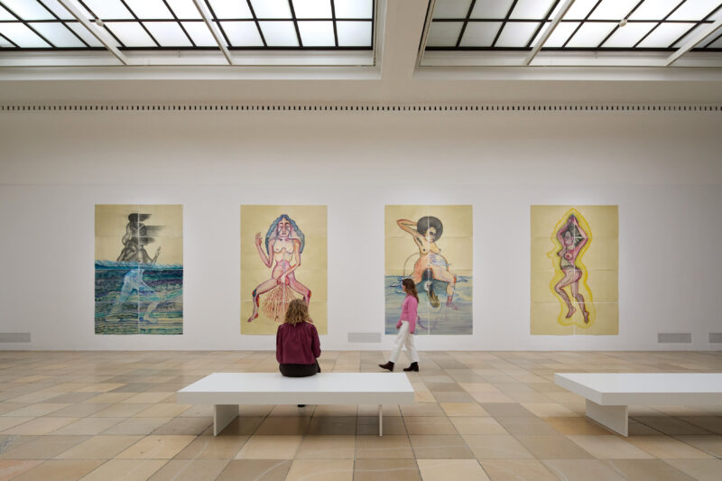 In einem Raum hängen bunte Bilder und eine Person sitz auf einer Bank davor.