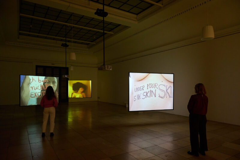 In einem dunklen Raum hängen drei Bildschirme von der Decke udn zwei Personen sehen sich die Videos darauf an.