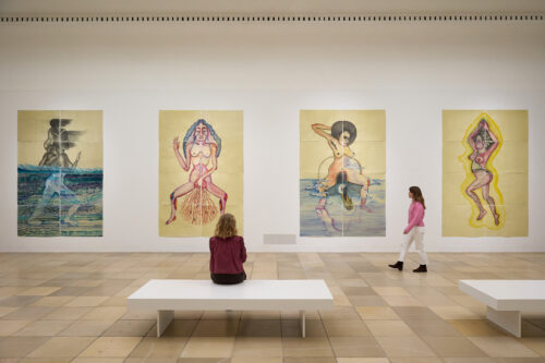 An einer Wand hängen Bilder. DAvor sitzt eine Person auf einer Bank und schaut sich diese an.