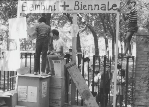 Ein schwarz- weiß Foto auf dem zwei Personen ein Plakat aufstellen auf dem steht: „Bambini + Biennale“