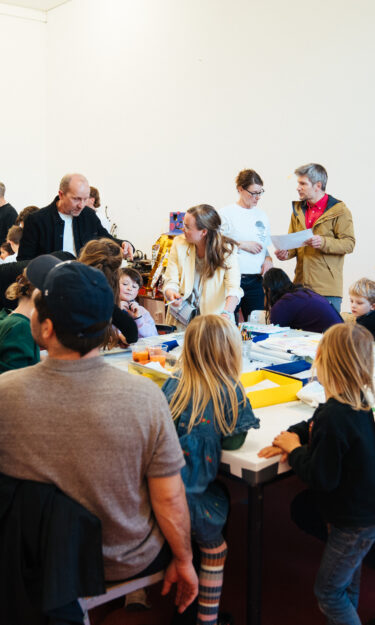 Open Haus meets Public Posession Photo Priscilla Grubo – Menschengruppe mit Kindern, die um einen Tisch stehen und sitzen.