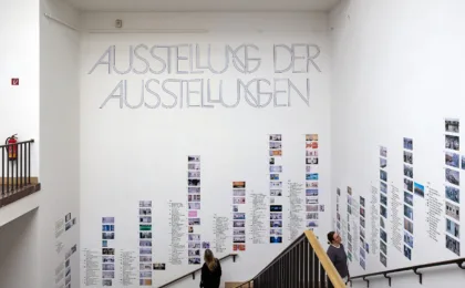 In einem Treppenaufgang sind  an den Wänden viele Kleine Bilder und der Schriftzug: „Ausstellung der Ausstellungen“ angebracht.