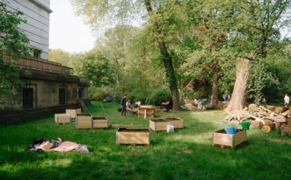 Auf einer grünen Wiese stehen Holzkisten mit Erde gefüllt. Die Sonne scheint und eine Person liegt auf einer Decke auf der Wiese und sonnt sich.