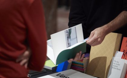 Ein Buch wird gerade aufgeschlagen. Im Hingtergrund befindet sich ein Tisch mit unterschiedlichen Büchern.