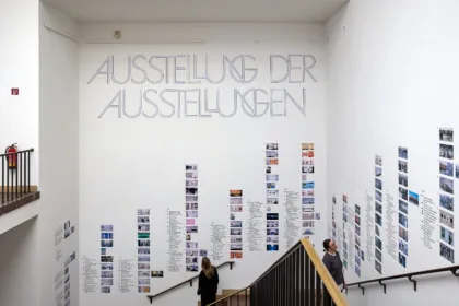 In einem Treppenaufgang sind  an den Wänden viele Kleine Bilder und der Schriftzug: „Ausstellung der Ausstellungen“ angebracht.