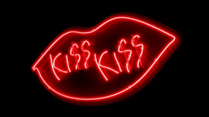 Auf schwarzem Grund befindet sich ein Kussmund aus roten Neonlichtröhren und der Schriftzug "KISS KISS".