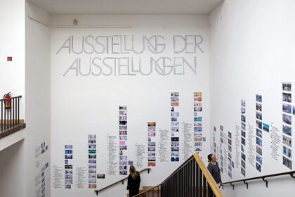 In einem Treppenaufgang sind  an den Wänden viele Kleine Bilder und der Schriftzug: „Ausstellung der Ausstellungen“ angebracht.