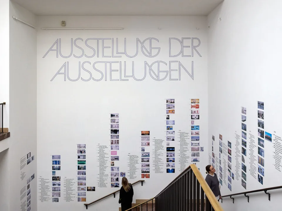 In einem Treppenaufgang sind  an den Wänden viele Kleine Bilder und der Schriftzug: „Ausstellung der Ausstellungen“ angebracht.