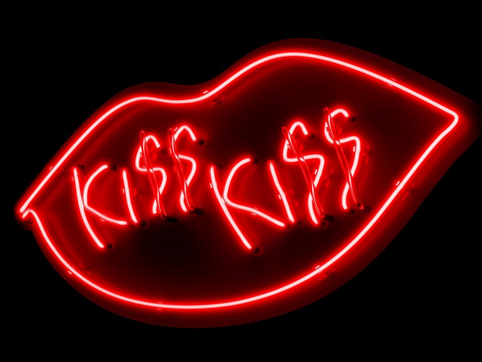 Auf schwarzem Grund befindet sich ein Kussmund aus roten Neonlichtröhren und der Schriftzug "KISS KISS".