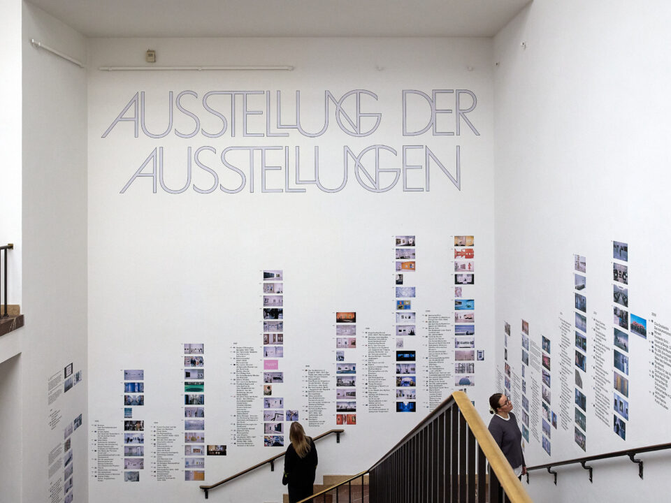 In einem Treppenaufgang sind  an den Wänden viele Kleine Bilder und der Schriftzug: „Ausstellung der Ausstellungen“ angebracht.