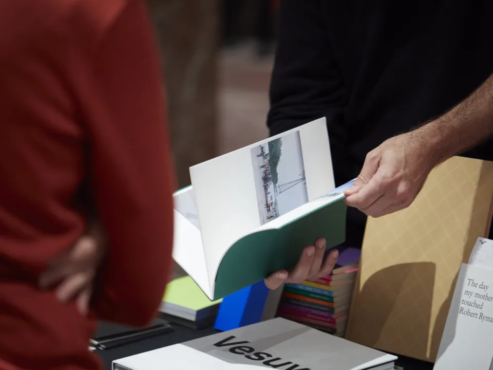 Ein Buch wird gerade aufgeschlagen. Im Hingtergrund befindet sich ein Tisch mit unterschiedlichen Büchern.