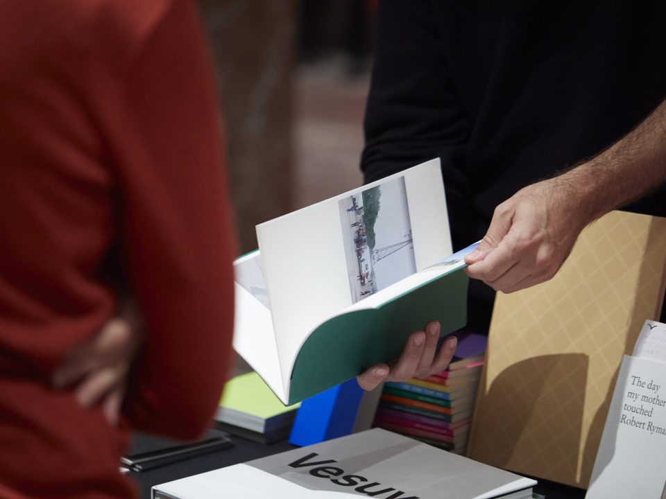 Ein Buch wird gerade aufgeschlagen. Im Hingtergrund befindet sich ein Tisch mit unterschiedlichen Büchern.