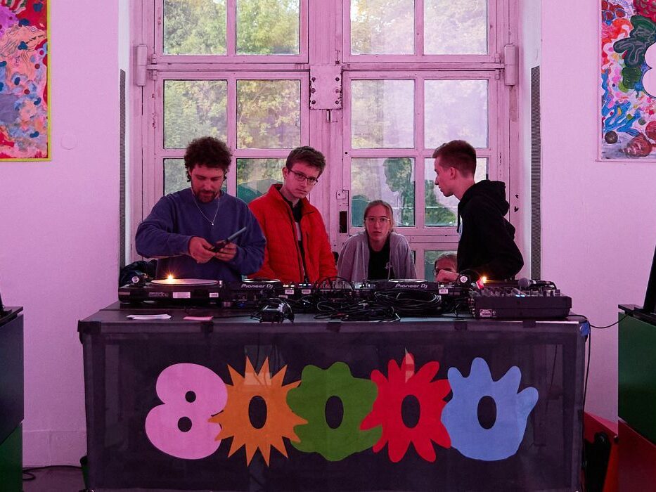 Mehrere Menschen stehen hinter einem DJ- Pult auf dem die Zahl 80000 angebracht ist.